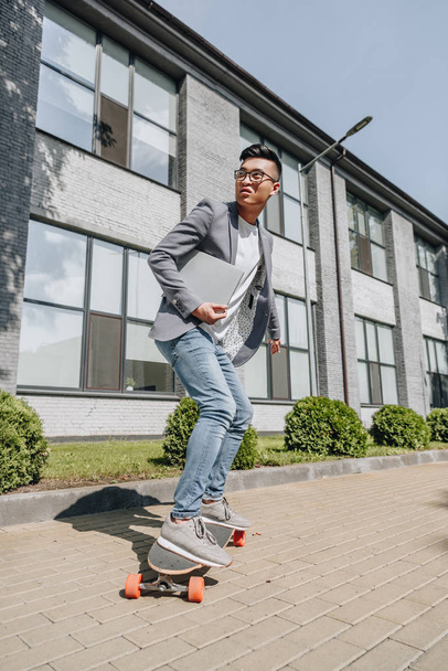 handsome asian man with laptop skateboarding on longboard - Foto, afbeelding