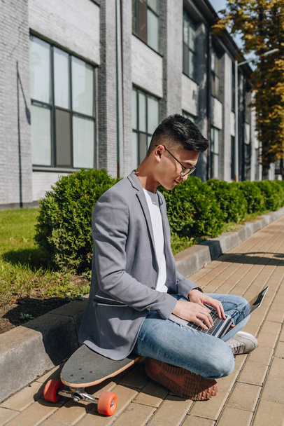 asian man typing on laptop while sitting on longboard on street - Foto, Imagen