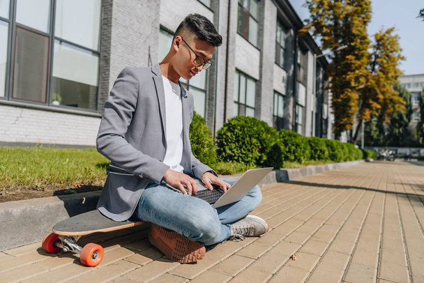 asian freelancer using laptop while sitting on longboard on street - Foto, Imagen