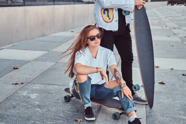 Hipster girl sitting on longboard while her boyfriend standing near outdoors. - Zdjęcie, obraz