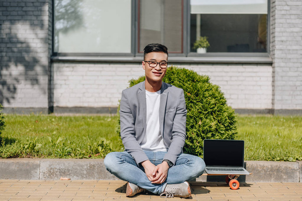 lächelnder asiatischer Mann sitzt auf Longboard mit Laptop mit leerem Bildschirm auf der Straße - Foto, Bild
