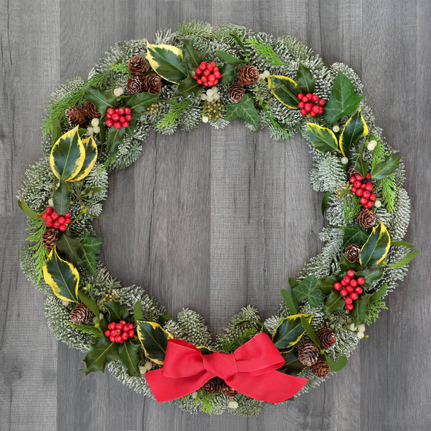 Christmas and winter wreath with holly, mistletoe, ivy, spruce fir, pine cones and a red bow on rustic grey wood  background. - Фото, изображение