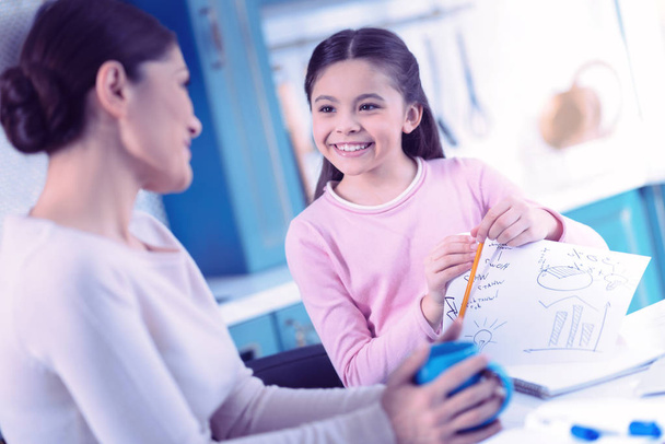 Menina positiva sorrindo para sua mãe enquanto fazia lição de casa com ela
 - Foto, Imagem