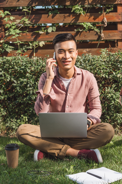 sonriente asiático hombre hablando en smartphone mientras remoto trabajo en portátil en parque
 - Foto, Imagen