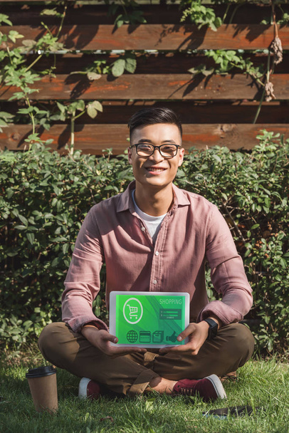 sourire homme asiatique assis sur herbe verte et montrant tablette avec panneau d'achat à l'écran dans les mains dans le parc
 - Photo, image