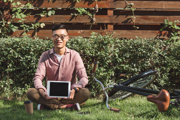 sonriente asiático hombre sentado en verde hierba y mostrando tableta con pantalla en blanco en las manos en parque
 - Foto, imagen