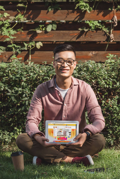 s úsměvem asijské muž sedí na zelené trávě a ukazuje tablet s aliexpress webové stránky na obrazovce v rukou v parku - Fotografie, Obrázek