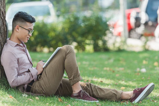 Widok z boku z azjatyckiej człowiek w okularach za pomocą cyfrowego tabletu na zielonej trawie w parku  - Zdjęcie, obraz