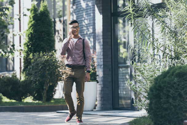  asian man with backpack having conversation on smartphone while walking on street - Φωτογραφία, εικόνα