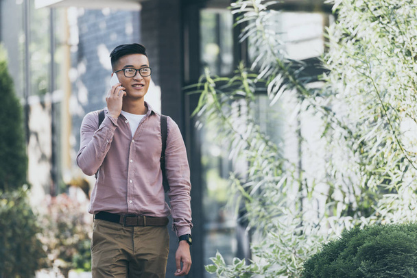 asiático hombre con mochila teniendo conversación en smartphone mientras caminando en calle
 - Foto, Imagen