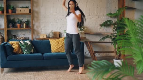 Joyful Asian girl is dancing on floor at home spinning and jumping listening to music in headphones using smartphone. Youth lifestyle and joy concept. - Filmagem, Vídeo
