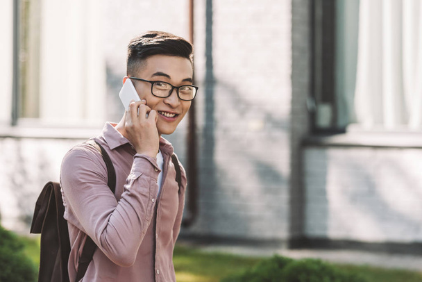  vista lateral de asiático joven en gafas teniendo conversación en smartphone en la calle
 - Foto, Imagen