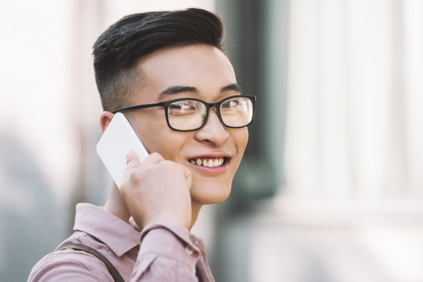  side view of smiling asian man in eyeglasses having conversation on smartphone on street - Photo, Image