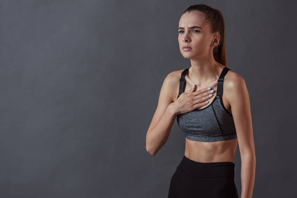 Beautiful girl in sportswear is touching her chest feeling pain, on gray background - Fotó, kép