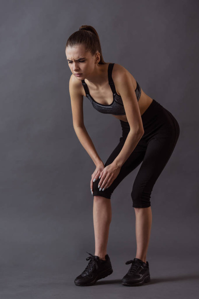 Beautiful girl in sportswear is touching her knee feeling pain, on gray background - Photo, Image