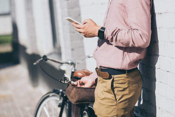 przycięte strzał człowieka w stylowe ubrania za pomocą smartfona o rower na ulicy - Zdjęcie, obraz