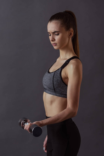 Beautiful girl in sportswear is doing exercises using dumbbell, on gray background - Photo, Image
