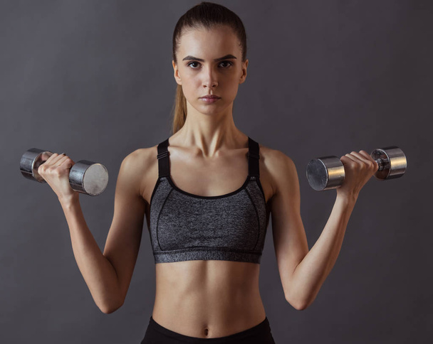Beautiful girl in sportswear is doing exercises using dumbbell and looking at camera, on gray background - Photo, Image