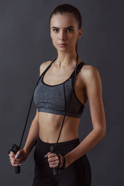 Beautiful girl in sportswear is holding a skipping rope and looking at camera, on gray background - Photo, Image