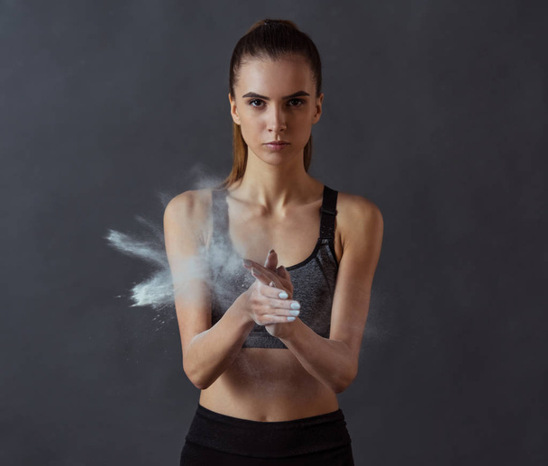 Beautiful girl in sportswear is using talcum powder for her hands and looking at camera, on gray background - Photo, Image