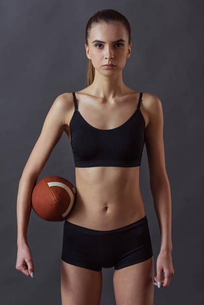 Beautiful girl in sportswear is holding an American football ball and looking at camera, on gray background - Zdjęcie, obraz