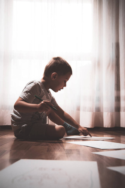 niño hace su tarea, escribe y dibuja en hojas de papel dispersas en el suelo
 - Foto, Imagen