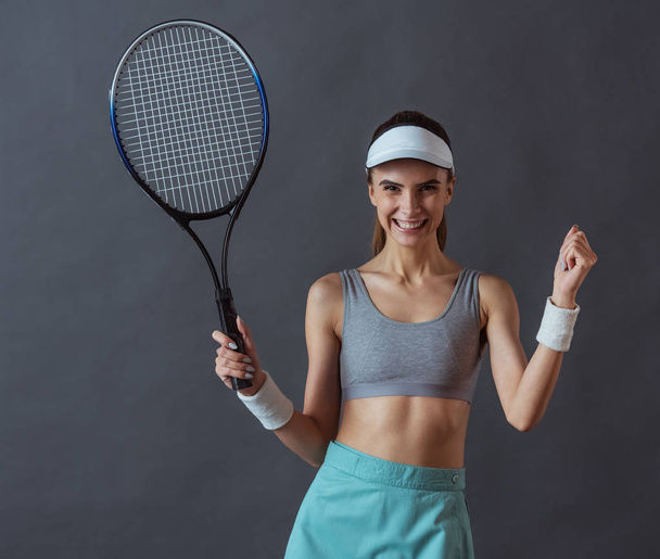schönes Mädchen in Sportbekleidung hält einen Tennisschläger in der Hand, blickt in die Kamera und lächelt, auf grauem Hintergrund - Foto, Bild