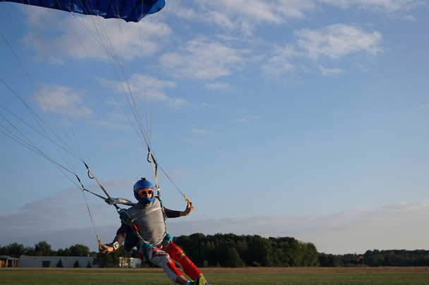 Paracaidista bajo un pequeño dosel azul oscuro de un paracaídas está aterrizando en el aeródromo, de cerca. Aterrizaje a alta velocidad de un paracaidista sobre el fondo de bosques y edificios
. - Foto, Imagen
