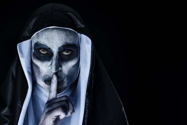Nahaufnahme einer beängstigend bösen Nonne, die ein typisches schwarz-weißes Gewand trägt und um Stille bittet, vor schwarzem Hintergrund mit einer Leerstelle rechts - Foto, Bild