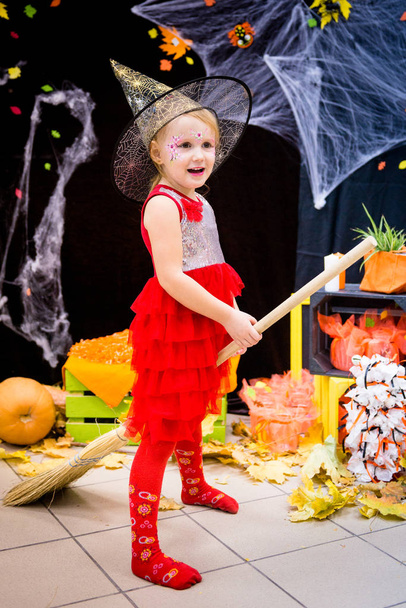 Un niño, una niña en forma de bruja en un palo de escoba, posa sobre el telón de fondo del paisaje de telarañas, calabazas y hojas de otoño en un día festivo de Halloween
. - Foto, Imagen