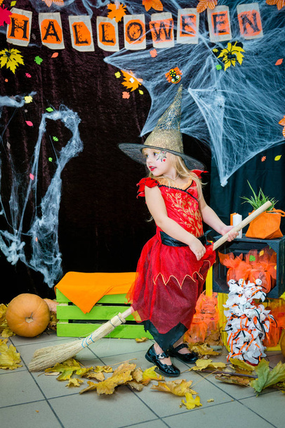 Un niño, una niña en forma de bruja en un palo de escoba, posa sobre el telón de fondo del paisaje de telarañas, calabazas y hojas de otoño en un día festivo de Halloween
. - Foto, imagen