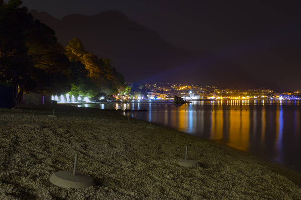 Night view of the illuminated seaside town of Baska Voda in Croatia. In the foreground is a pebble beach with the stands for parasols. - Photo, Image
