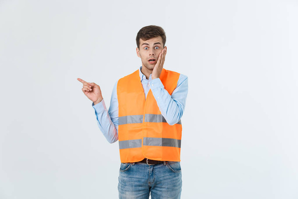 Handsome young professional engineer man over grey wall wearing orange vest amazed and surprised pointing with fingers. - Photo, Image