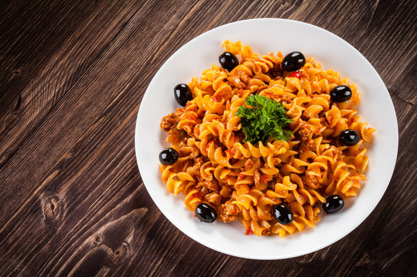 Spiral pasta with bolognese sauce, black olives and parsley on white plate on wooden table - 写真・画像