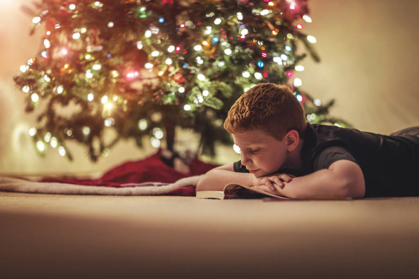 Young boy reading a book under the Christmas tree with added vintage filter and soft focus - Foto, Imagem