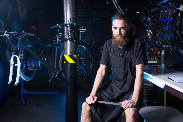 Portrait of small business owner of young man with beard. Guy bicycle mechanic workshop worker sitting with tool in his hand in a working black clothes in an apron in the background of a bicycle shop. - Photo, Image