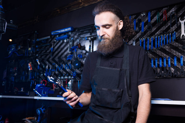Retrato del dueño de una pequeña empresa de un joven con barba. Guy mecánico de bicicleta trabajador de taller sentado con la herramienta en la mano en una ropa de trabajo negro en un delantal en el fondo de una tienda de bicicletas
. - Foto, imagen