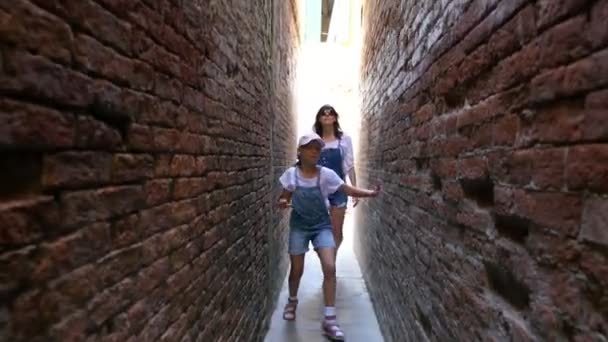 VENICE, ITALY - JULY 7, 2018: along very narrow street of Venice, between old houses, a teenager girl, child and young woman walk and dance, in shorts and hats. summer hot day. - Felvétel, videó