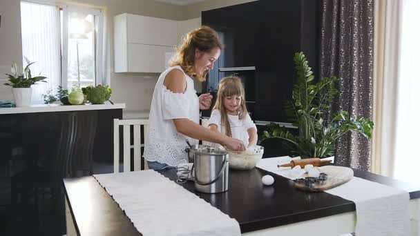Ride the camera. A young woman and little daughter are baking in kitchen. Young mom and her little girl is kneading a dough together in the big glass bowl in the kitchen - Filmagem, Vídeo