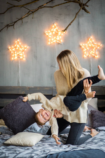 Mother and little boy are having fun on cosy bed in front of festivaly decorated wall with shining stars. Cute boy is laughing. New Year`s celebration. - Фото, изображение