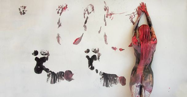 Молодая художественно абстрактно окрашенная женщина с черно-красно-белым цветом, краска, валит перед белой стеной в студии и оставляет красочные цветные отпечатки
. - Фото, изображение