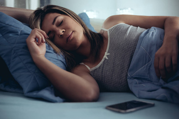 技術と人のコンセプト - 夜に自宅のベッドで眠っているスマート フォンを持つ若い女性 - 写真・画像