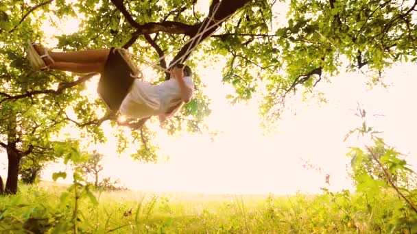 mooi meisje swingen op schommel verlicht door de stralen van de zon. Rope swing op boomtakken in het park. Slow motion. - Video