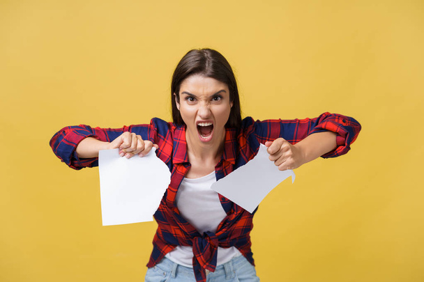 Aggressive young caucasian woman tears contract or white sheet of paper. Isolated over yellow background - Foto, Imagen