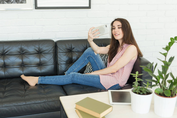 Conceito de tecnologia, beleza e pessoas - jovem mulher sorridente tentando fazer foto usando smartphone e deitada no sofá em casa tocando seu cabelo
. - Foto, Imagem
