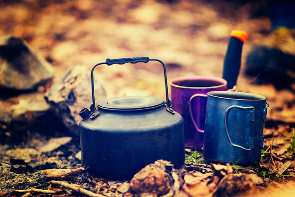 Small steel kettle stands next to  two cups on the land in the autumn forest on blurry warm background. Waiting for coffee. Vintage image. - 写真・画像
