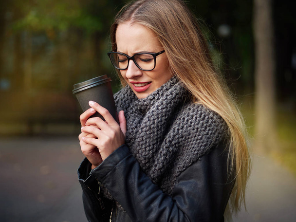 Portrait of a beautiful stylish young blonde with make-up in black glasses a leather jacket and a knitted scarf. Frozen girl drinks hot drink in a black mug in the autumn cold park - Foto, Bild