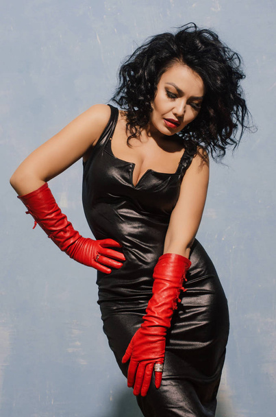 Prachtig fetish model dragen van zwarte spandex jurk en lange rode lederen handschoenen poseren in de buurt van blauwe muur. - Foto, afbeelding
