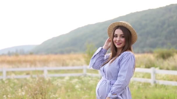 Беременная женщина в природе примеряет вязаную шляпу. Фон поля и белый забор возле фермы. горы
 - Кадры, видео
