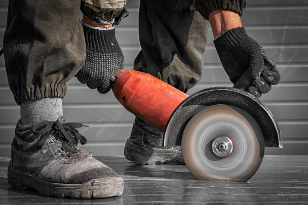 Un soldador joven en un conjunto de trabajo y guantes de trabajo muele un objeto de metal con una amoladora angular en el lado de la restricción
 - Foto, Imagen